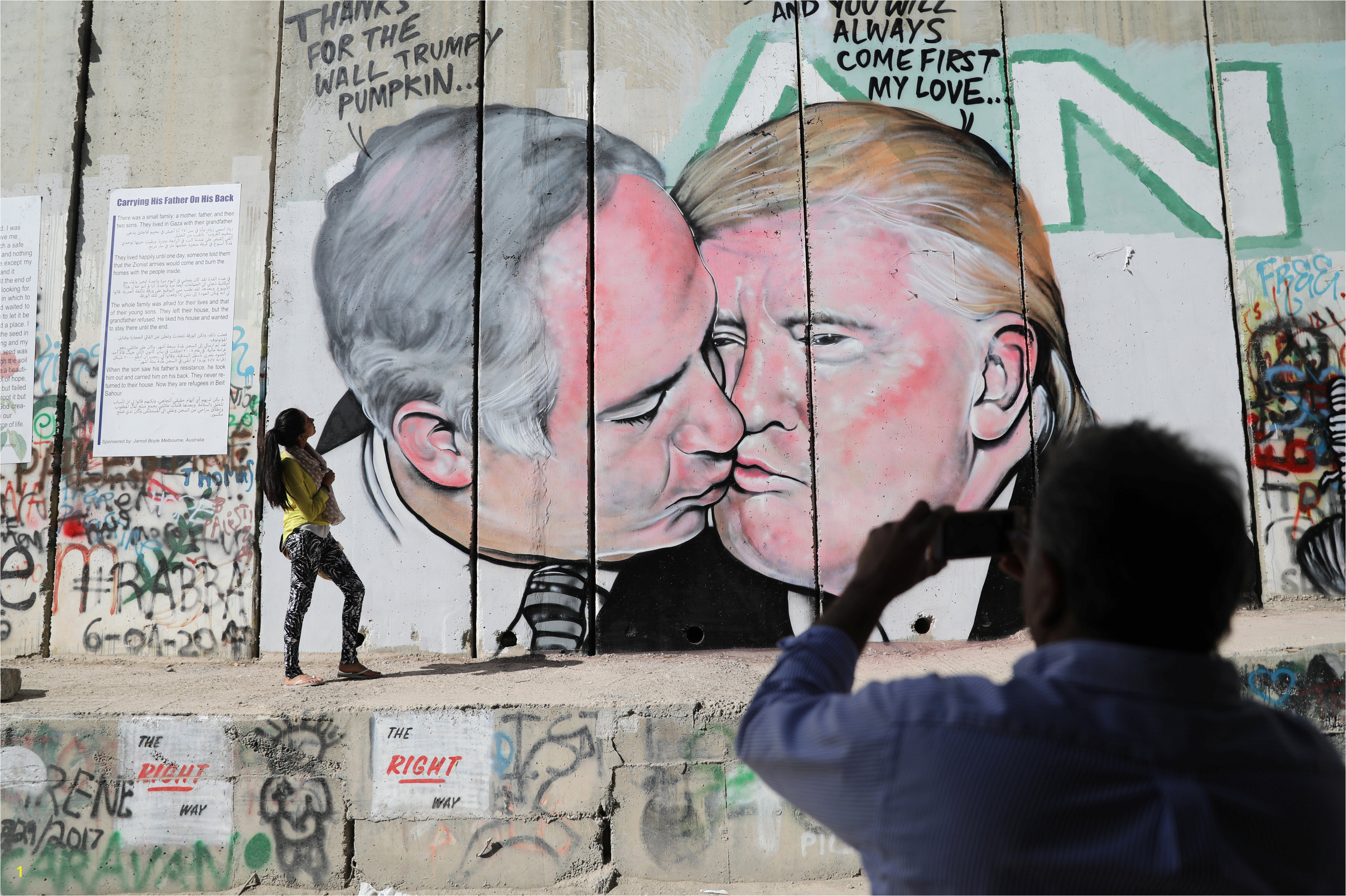 A man photographs a woman as she stands next to a mural depicting U S President Donald